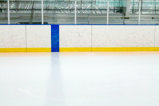 Photograph from the center of the ice at a ice hockey rink.  The blue line is on the boards and in the ice of the indoor arena.  Through the glass, metal bleachers can be seen in this small town ice rink.  The boards are covered with black marks from pucks and is lined with a yellow stripe.