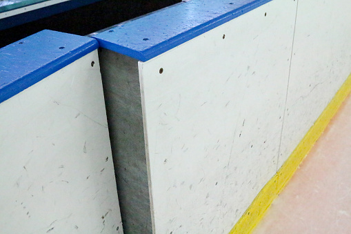 The door through the boards at an ice hockey rink is partially open to the player's bench.  The stadium boards are white with blue on top and a yellow line near the ice.  The slightly open door could represent a player going onto or getting off of the bench.