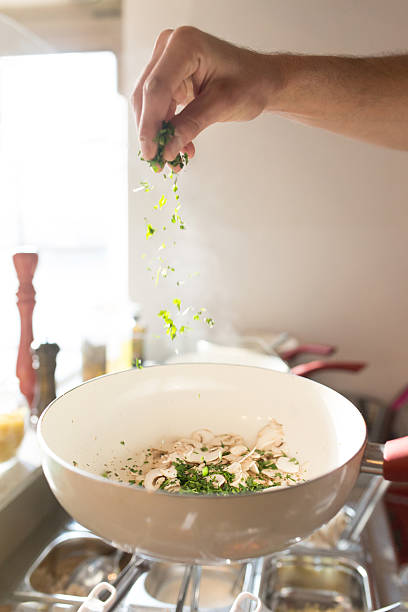 Human hand spicing champignon mushrooms with parsley and basil in a cooking pan.