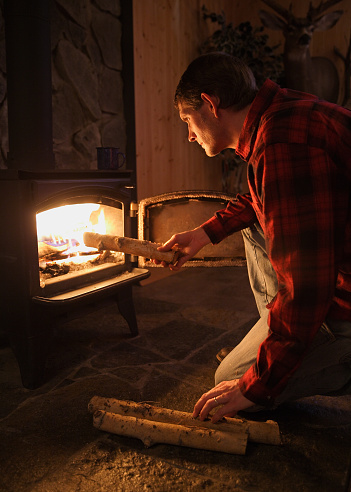 mid-adult man adding logs to a wood burning stove fire in rustic setting