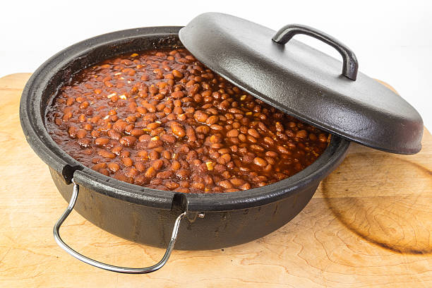 Southern Style Baked Beans Large Cast Iron Dutch Oven filled with piping hot Southern Style Sweet Baked Beans resting on old wooden cutting board with white background. baked beans stock pictures, royalty-free photos & images