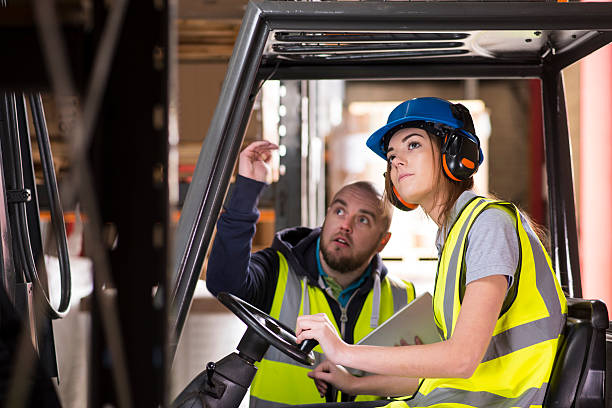 female forklift driver training A young female forklift driver is being trained on approaching the racking of the warehouse . A male colleague or trainer can be seen advising her on how to perform the manoeuvre . She is wearing a hi vis jacket , hard hat and ear defenders . ear protectors stock pictures, royalty-free photos & images