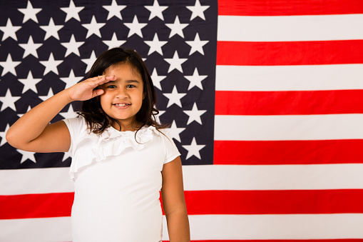 Color photo of a happy, young girl saluting America and our troops in front of a large American flag. Composed with room for text on right.
