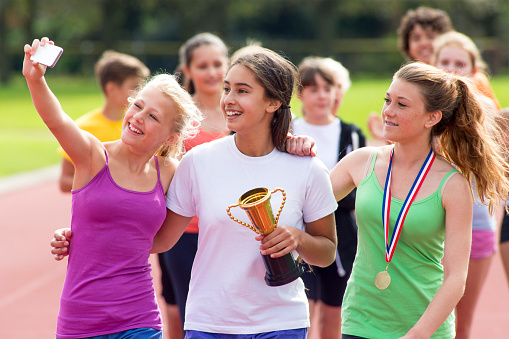 Three girls taking a selfie with their newly received medals and trophies after winning and placing in a track race.