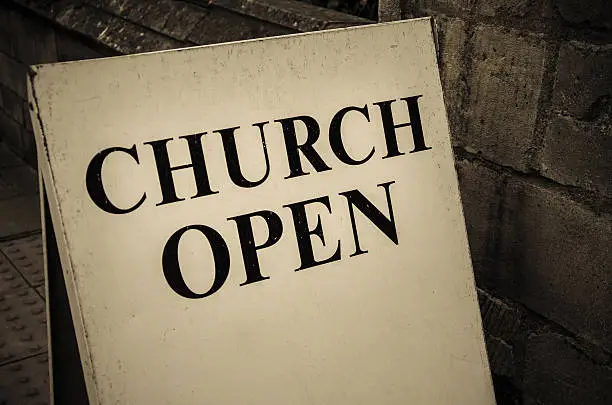 A sign at a church entrance indicating it is open to public and welcoming people inside. The text is on capital Although the image is in colour, it is grey toned.