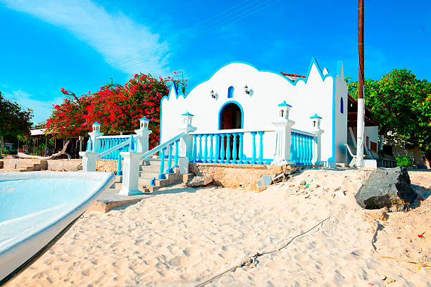 Digital photograph of beautiful white and blue church located in Los Roques, a tropical Island off the coast line of Venezuela, during a bright and warm day. The church is feet away from the beachline and surrounded by tropical vegetation and sand. The architecture of it is very island-like and blends in with the surroundings seamingless