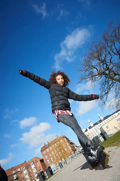 Full figure image of a cheerful teenage girl skating in an urban scene with buildings in the background. The girl is of mixed race with dark skin and brown, curly hair. The image is photographed from a low angle view.