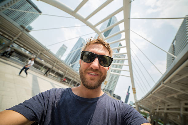 Young cheerful tourist man in Bangkok taking a selfie on the Tha Sathorn place station going to the sky train. Offices buildings and towers on the background. The man is wearing casual clothing and sunglasses in summer, people walking around.