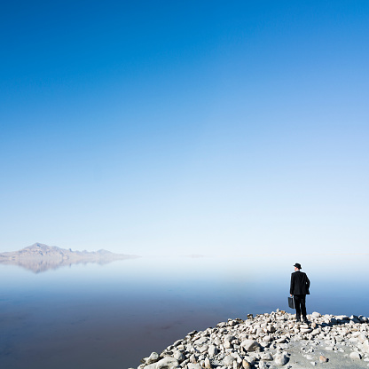 Businessman standing in the front of vast open space looking into a new land on the horizon in search of new business opportunity, plenty of copyspace, blue clear sky and calm waters ahead of him. 