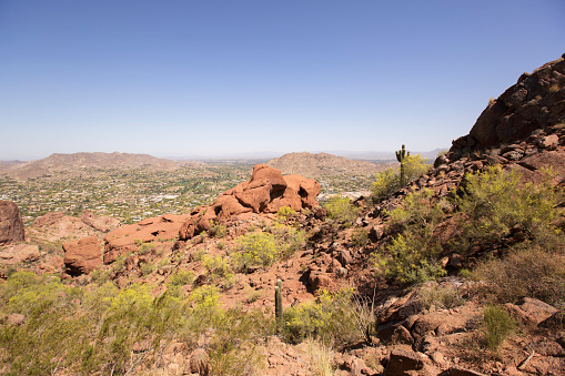 This image is taken while climbing Camelback Mountain in Arizona. The rocky terrain overlooks Scottsdale. Landscape scenery is speckled with cactus and small shrubs. The sky is blue with hot sunshine. Image taken in April.