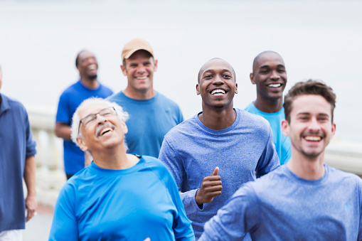 A multi-ethnic group of men wearing casual blue shirts, running or power walking together.  The focus is on the young African American man in his late 20s, in the middle, looking at the camera.  They are together supporting a common cause, perhaps cancer awareness.