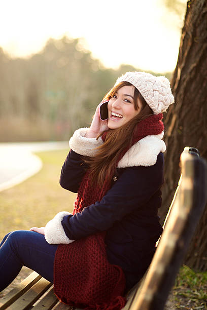 Smiling girl talking on mobile phone on bench in park in the Winter