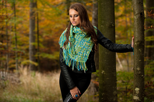 Pretty young woman with long brown hair on a walk in forest on late autumn day in fashoinable dress with scarf. She is standing, with one hand leaning against a tree and looking into floor.