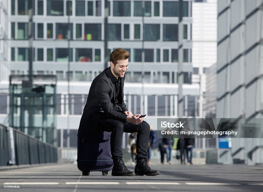 Cheerful young man sitting on suitcase looking at mobile phone Portrait of a cheerful young man sitting on suitcase at station looking at mobile phone 20-29 Years Stock Photo