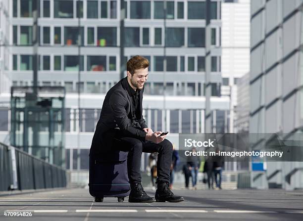 Cheerful Young Man Sitting On Suitcase Looking At Mobile Phone Stock Photo - Download Image Now