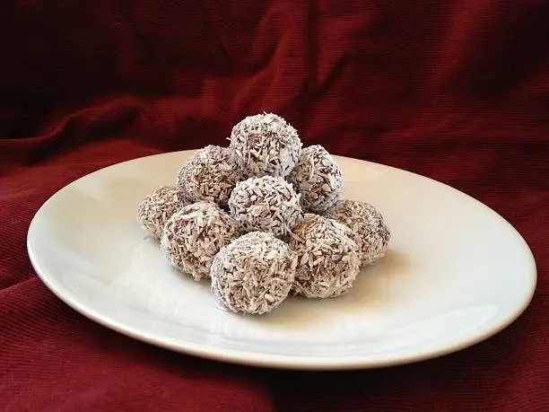 Swedish chocolate balls (chokladbollar) with coconut flakes on a white plate with a crimson background.