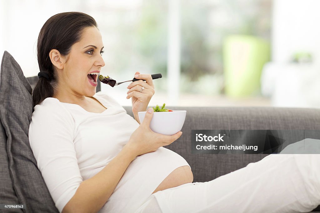 pregnant woman eating vegetables on sofa attractive pregnant woman eating vegetables on sofa Abdomen Stock Photo
