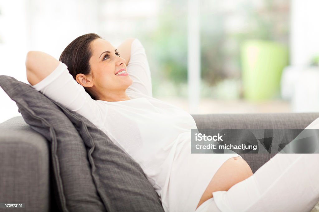 Smiling pregnant woman leaning against side arm of couch happy pregnant woman lying on couch daydreaming Abdomen Stock Photo