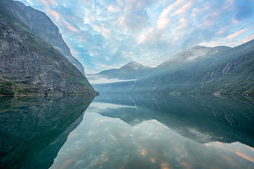 Beautiful bracketed HDR shot of the famed Geirangerfjord in Norway from a Cruise ship in the early morning predawn hours with a great dramatic sky above and very calm ocean water.
