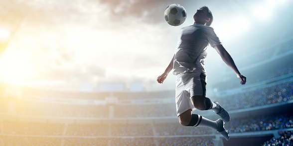 A male soccer player makes a dramatic play by jumping vertically. He attempts to hit the ball with. The stadium is blurred behind him. Only the lights of the stadium shine brightly, creating a halo effect around the bulbs.