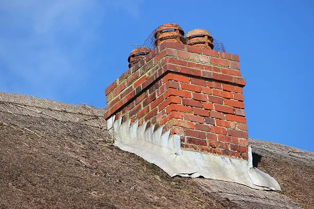 Photo showing the close-up detail of a thatched roof, with chimney pots and a red-brick chimney stack, which is made waterproof by lead flashing around its base.  The chimney pots are covered with chicken wire, to stop debris and birds falling down.