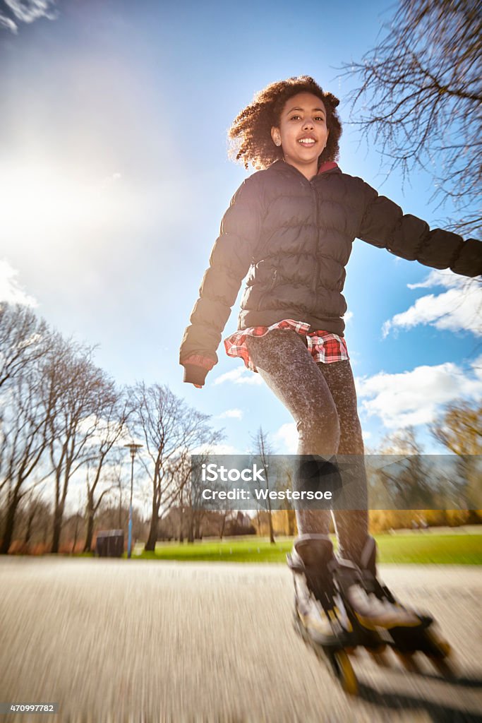 Roller skating tenage girl in motion, backlit urban scene outdoor Full figure image of a cheerful teenage girl skating in a park with trees and skies in the background. The girl is of mixed race with dark skin and brown, curly hair. The image is photographed from a low angle view and has a vintage look with some motion blur. Teenage Girls Stock Photo