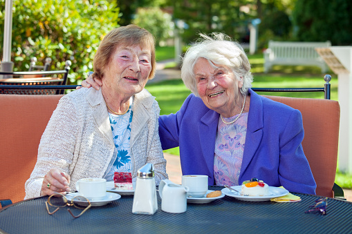 Two Happy Senior Women Relaxing at the Garden Table with Cups of Coffee and Snacks  Looking at the Camera.