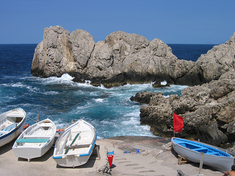 This is a scene from Faro Beach on the island of Capri in Italy.  Three white boats and one blue boat, as well as a red flag are part of this beach of cliffs and raging sea.