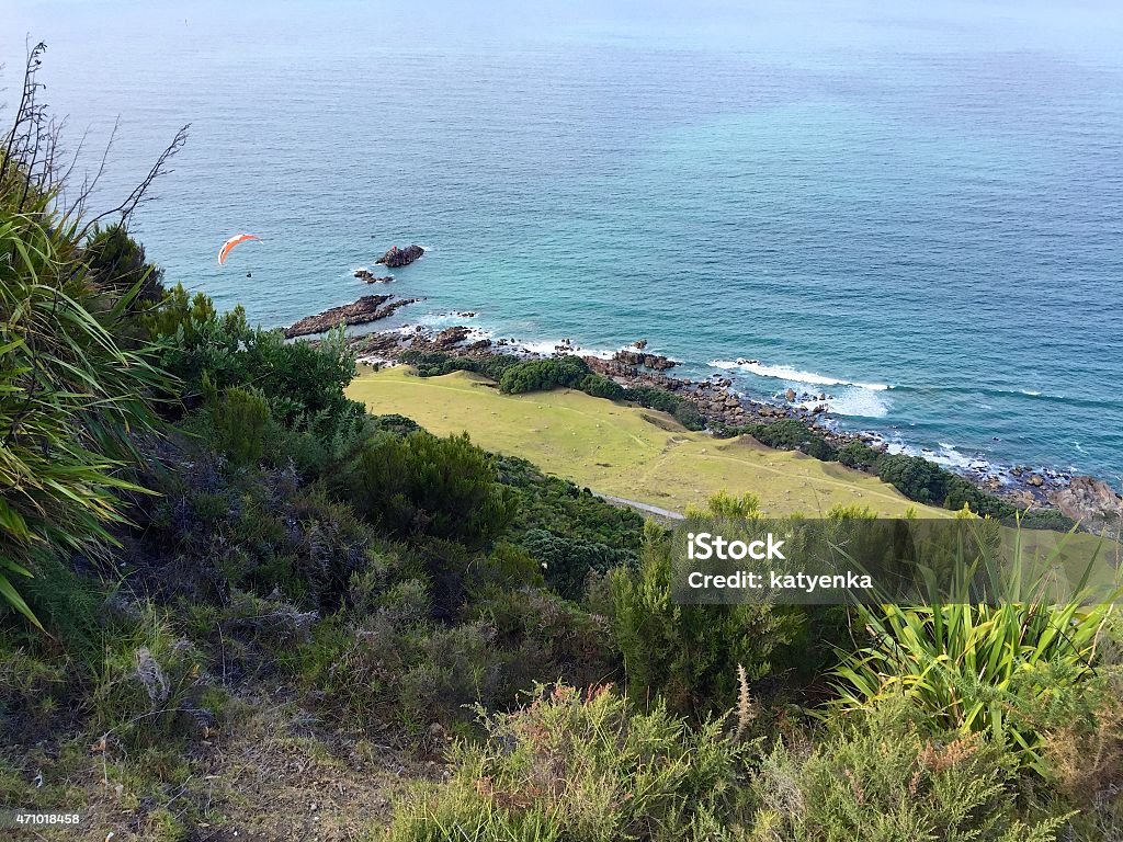 Paraglider over the Pacific Ocean in Mount Maunganui, New Zealand A paraglider is seen in the distance below gliding toward the water in Mount Maunganui, New Zealand. The view from above is taken from Mauao, looking down at the Pacific Ocean and the waves breaking on shore. Summertime. 2015 Stock Photo