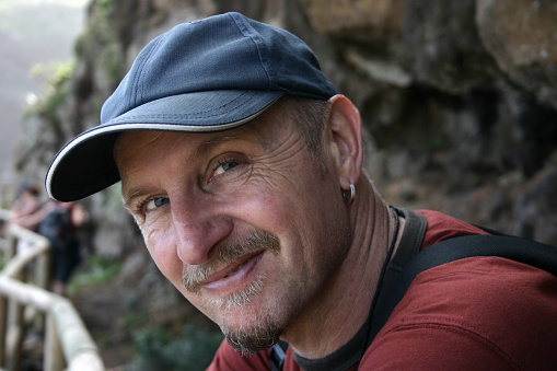 Head and shoulders of male hiker looking sideways into the camera with a baseball hat on.