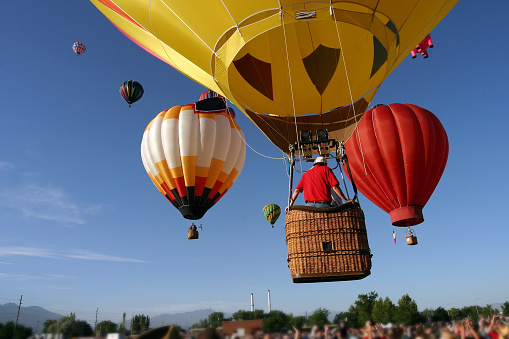 Several brightly colored hot air balloons take to the sky as a crowd of people looks on (faces are blurred to protect the \