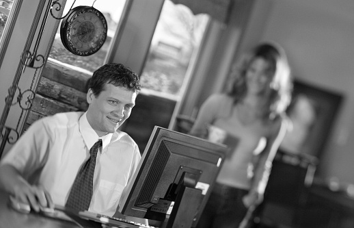 Young man working at the desk on the computer, lady in the background out of focus.