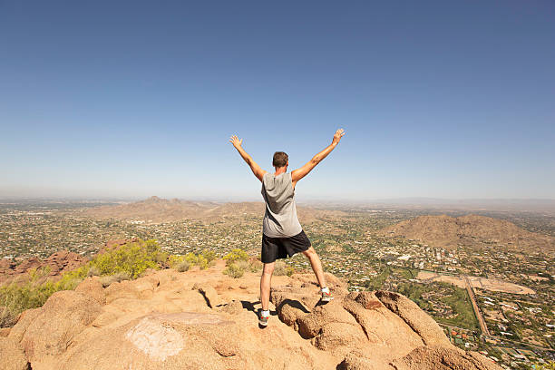 Man conquering mountain climb - leadership This image is taken while climbing to the top of Camelback Mountain in Arizona. The rocky terrain overlooks Scottsdale. Landscape scenery is speckled with cactus and small shrubs. The sky is blue with hot sunshine. Image taken in April. Image can be used as a leadership analogy of achieving success with determination through challenging experiences. sleeveless top stock pictures, royalty-free photos & images