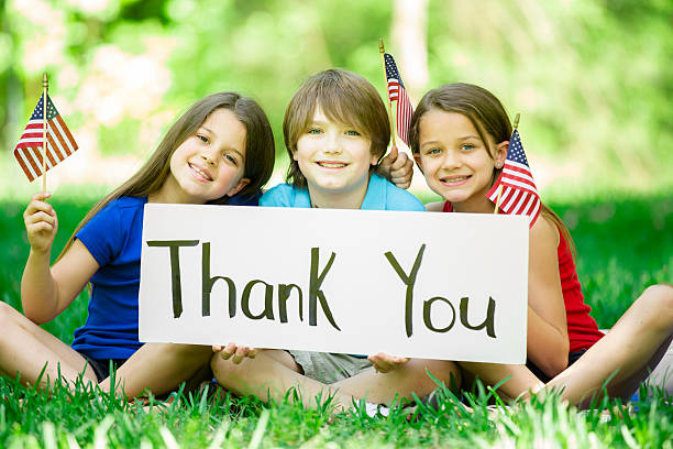Children hold "Thank You" sign with American flags. Memorial Day. Three American children hold "Thank You" sign outdoors in the spring or summer season in honor of US veterans...commemorating the USA Memorial Day, July 4th, or Veteran's Day holidays.  The three siblings have big smiles and hold American flags. Two little girls and one little boy are all elementary ages. Beautiful nature background. thank you veterans day stock pictures, royalty-free photos & images