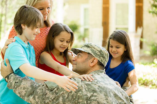U.S. military veteran welcomed home by family. Mom, children. Home. Handsome U.S.A. military soldier being welcomed home by his family. Wife, two little girls and one boy children. Home in background. He wears his military uniform and is excited to see his family. The children and wife eagerly give him a big hug. Thank you veterans for your service to your country! Memorial Day, Veteran's Day. proud army mom stock pictures, royalty-free photos & images