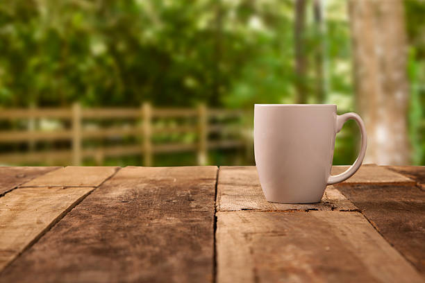 Composite image. Rustic, weathered wooden table with a white coffee cup on top of it.  Beautiful spring or summer background with a wooden fence. Sunny forest area, park, or backyard outdoor space. No people in this tranquil morning scene.
