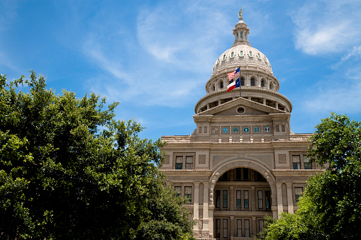 capitol in austin, texas, a monumental building with the us and the texan flag between large green summer trees.