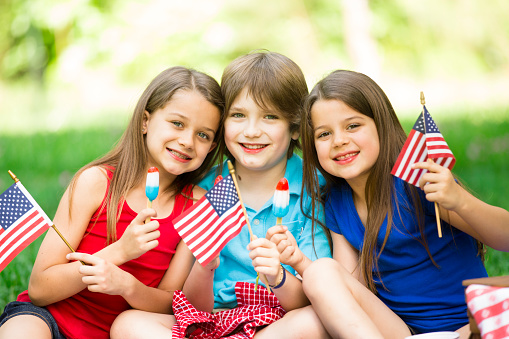 Three American children enjoy a summer picnic outdoors to celebrate USA's Memorial Day, Independence Day, or Labor Day holiday.  The three siblings enjoy yummy red, white, and blue popsicles. Two little girls and one little boy are all elementary ages. Beautiful nature background. American flags.
