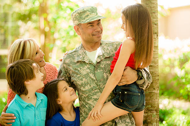 American military veteran welcomed home by family. Mom, children. Home. Handsome U.S.A. military soldier being welcomed home by his family. Wife, two little girls and one boy children. Home in background. He wears his military uniform and is excited to see his family. The children and wife eagerly give him a big hug. Thank you veterans for your service to your country! Memorial Day, Veteran's Day. proud army mom stock pictures, royalty-free photos & images