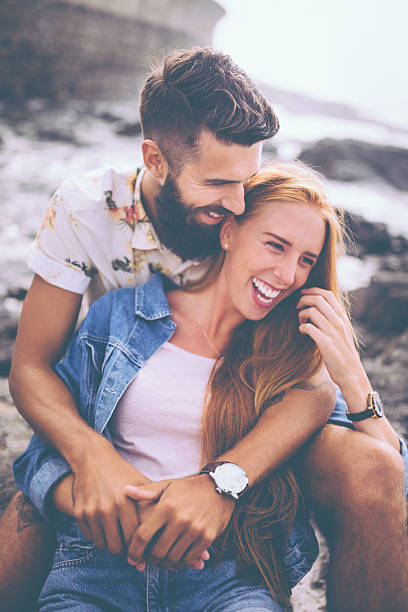 retro pareja riendo juntos mientras se distiende en rocks at seaside - couple old fashioned hipster holding hands fotografías e imágenes de stock