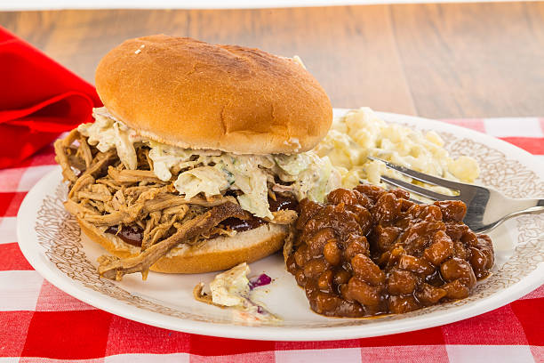 Memphis Barbecue Sandwich Pulled Pork on Bun with barbecue sauce and coleslaw.  Served with southern style baked beans and potato salad on picnic table with red plaid tablecloth. baked beans stock pictures, royalty-free photos & images