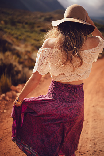 Rearview of a boho girl wearing a purple skirt walking down a dirt road on a summer day