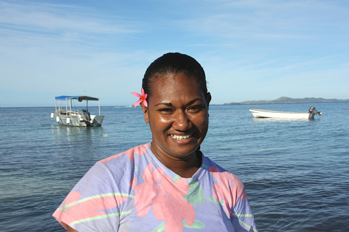 Gracious young native of the Fiji islands smiles broadly at the camera; South Pacific ocean and two small boats in the background. 