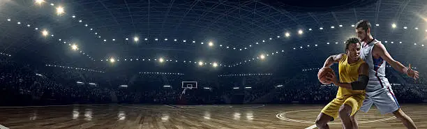 Low angle panoramic view of a professional basketball game. A player failed to block the opposite team player with a ball. A game is in a indoor floodlit basketball arena. All players are wearing generic unbranded basketball uniform.
