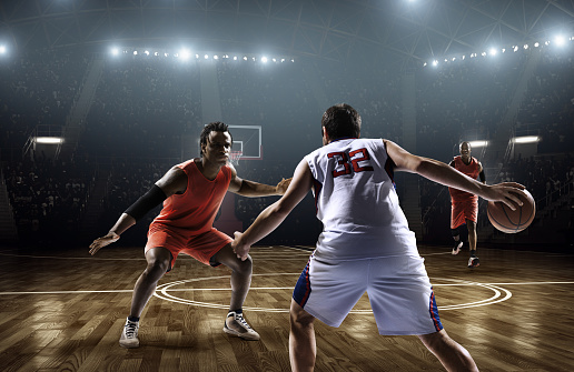 Image from a low angle viewpoint of a basketball player bribling basketball in front of rival players. The action takes place in generic indoor floodlit basketball arena full of spectators. All players are wearing generic unbranded basketball uniform.