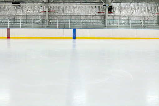 Photograph across the empty ice at an indoor hockey rink.  Through the glass, metal bleachers can be seen in this small town hockey arena.  The camera is lined up with the blue line, which is through the ice and on the boards.  The red center line can also be seen.  The ice reflects the lights.