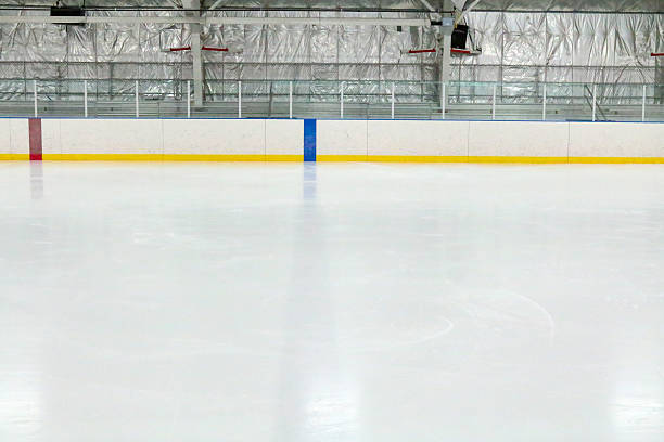 View across the empty ice at an indoor hockey rink Photograph across the empty ice at an indoor hockey rink.  Through the glass, metal bleachers can be seen in this small town hockey arena.  The camera is lined up with the blue line, which is through the ice and on the boards.  The red center line can also be seen.  The ice reflects the lights. ice rink stock pictures, royalty-free photos & images