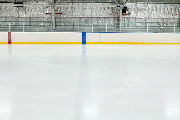 View across the empty ice at an indoor hockey rink Photograph across the empty ice at an indoor hockey rink.  Through the glass, metal bleachers can be seen in this small town hockey arena.  The camera is lined up with the blue line, which is through the ice and on the boards.  The red center line can also be seen.  The ice reflects the lights. skate rink stock pictures, royalty-free photos & images