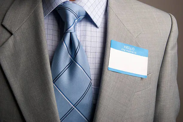Close up of simple blue tie with Windsor knot against checkered shirt and sober gray suit, balanced by a "Hello My Name Is" tag