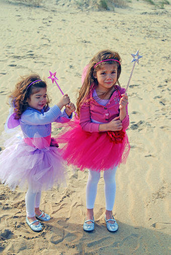 Two young girls dressed as fairies complet with wands stand crying and sad on a beach.